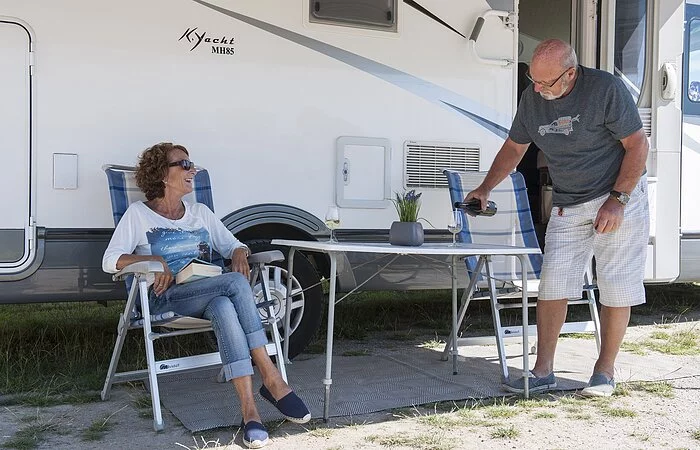 Tisch mit zwei Stühlen und zwei Personen vor einem Wohnmobil beim Weintrinken