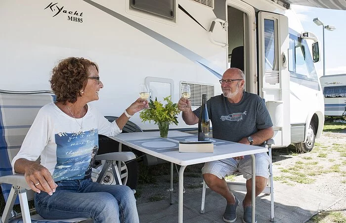 Zwei Personen sitzen vor dem Wohnmobil am Tisch und stoßen mit Wein an
