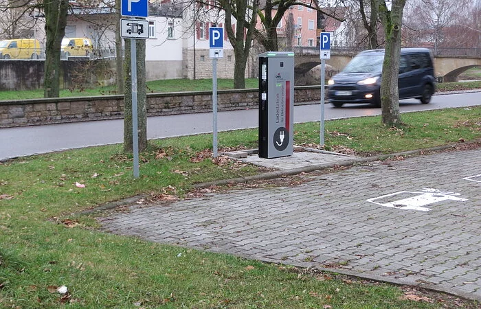 Beschilderung für Wohnmobilstellplatz sowie für Pkw(s). Nebendran eine Ladestation für Elektrofahrzeuge. Im Hintergrund Wohnhäuser und eine alte Steinbrücke