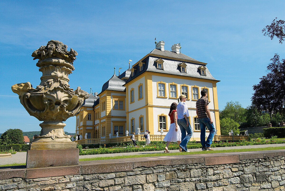 Mauer mit großer vasenähnlicher Skulptur auf einer Ecke, im Hintergrund barockes Schloss
