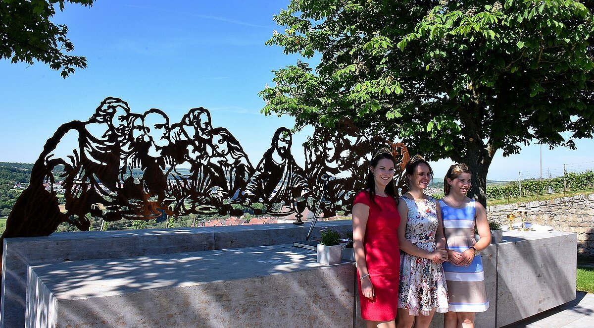 kleine Menschengruppe vor einer Skulptur mit Bank und Bäumen rechts und links. Die Skulptur stellt filigran mehrere Menschen im Umriss dar