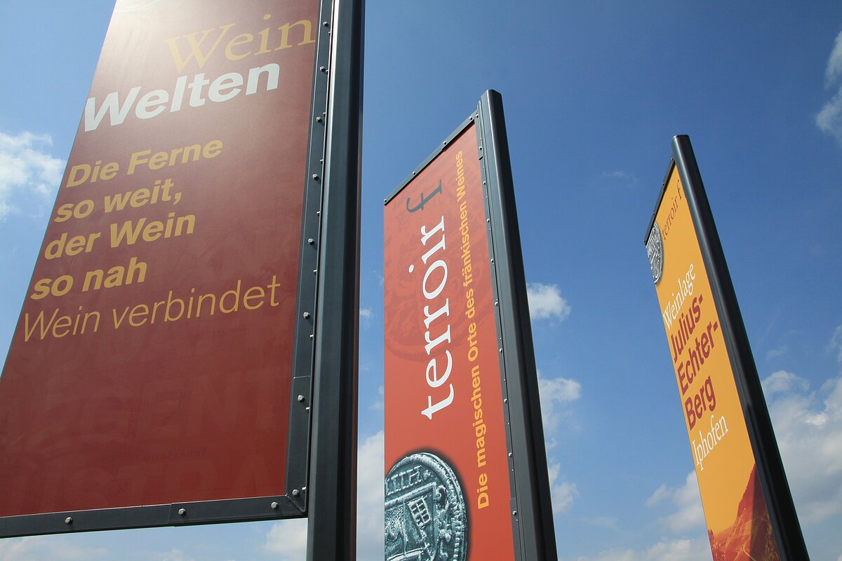 Drei beschriftete, rot, orange und gelbe,  rigide Fahnen gen Himmel