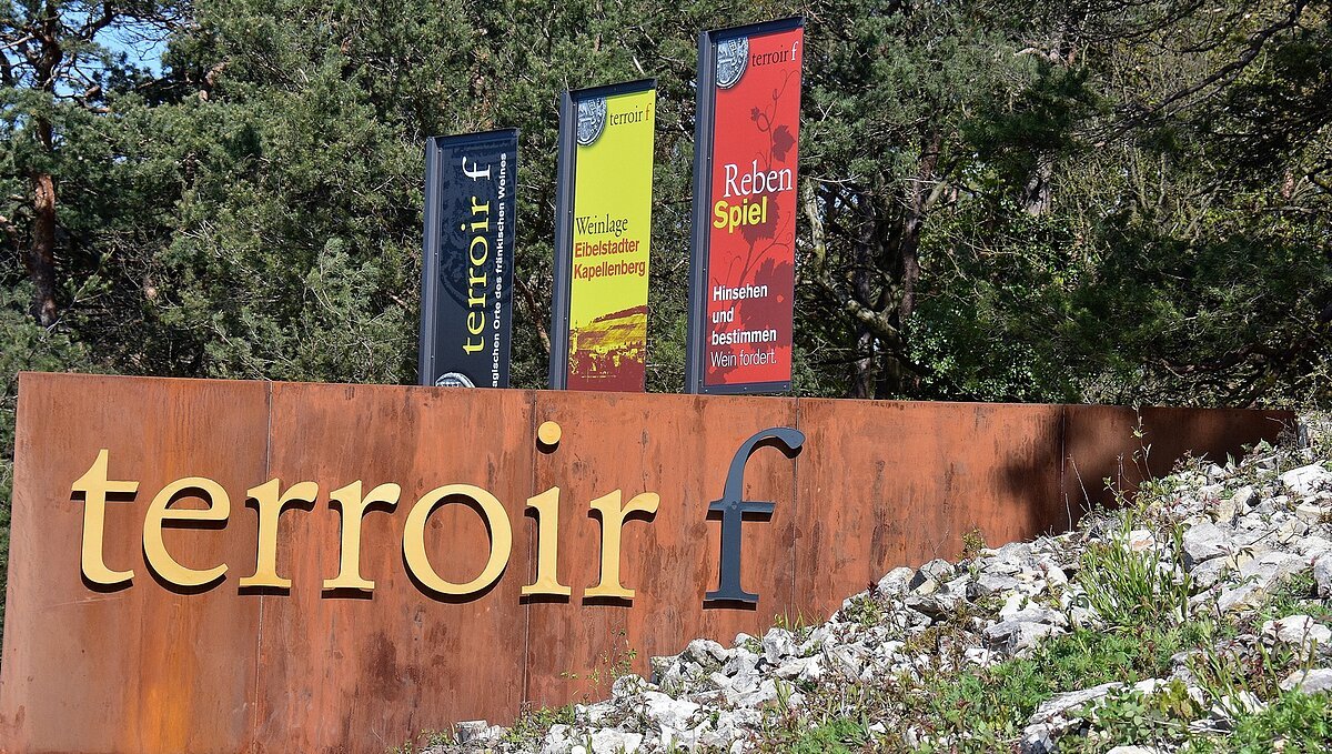Stahlwand mit Aufschrift terroir f, rechts Steinhügel, darüber mittig 3 farbige Fahnen mit Aufschrift, im Hintergrund Bäume