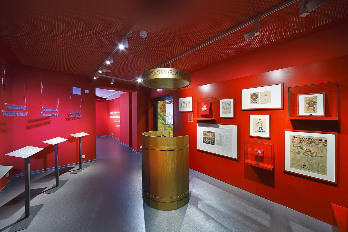 Roter Innenraum, grauer Boden, an der Wand rechts Bilder und Ausstellungsstücke, links Aufsteller in weiß, in der Mitte ein Säule