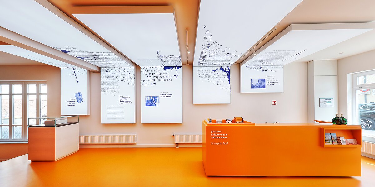 Heller Ausstellungsraum von innen, ohne Menschen, mit Fenstern und orangenen Möbeln