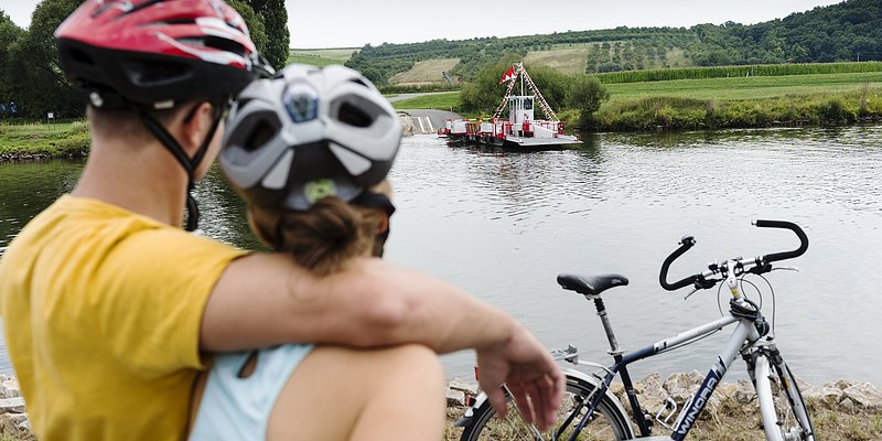 Ein Mann und eine Frau mit Sturzhelmen und Rädern blicken von der Fähre in die Landschaft